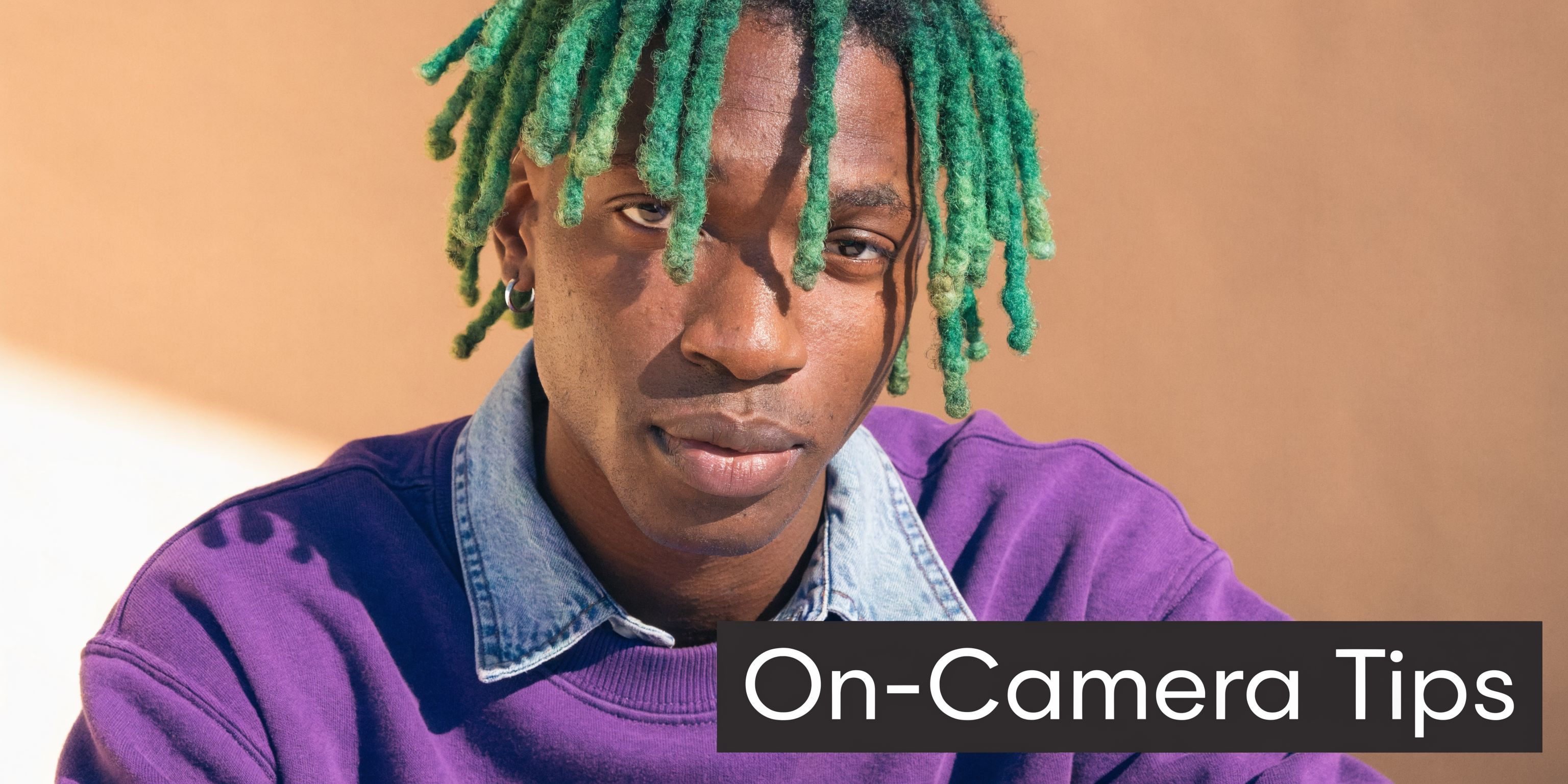 A young Black man with vibrant green dreadlocks wearing a purple sweatshirt looks directly at the camera.