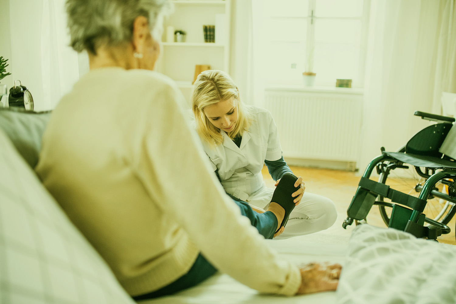 Pflegekraft untersucht den Fuß einer älteren Frau, die auf einem Bett sitzt, neben einem Rollstuhl im hellen Zimmer.
