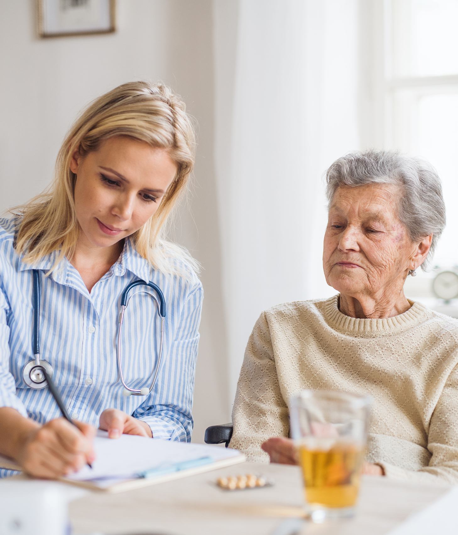 Junge Ärztin mit Stethoskop schreibt Notizen, während eine ältere Frau mit Pullover daneben sitzt.
