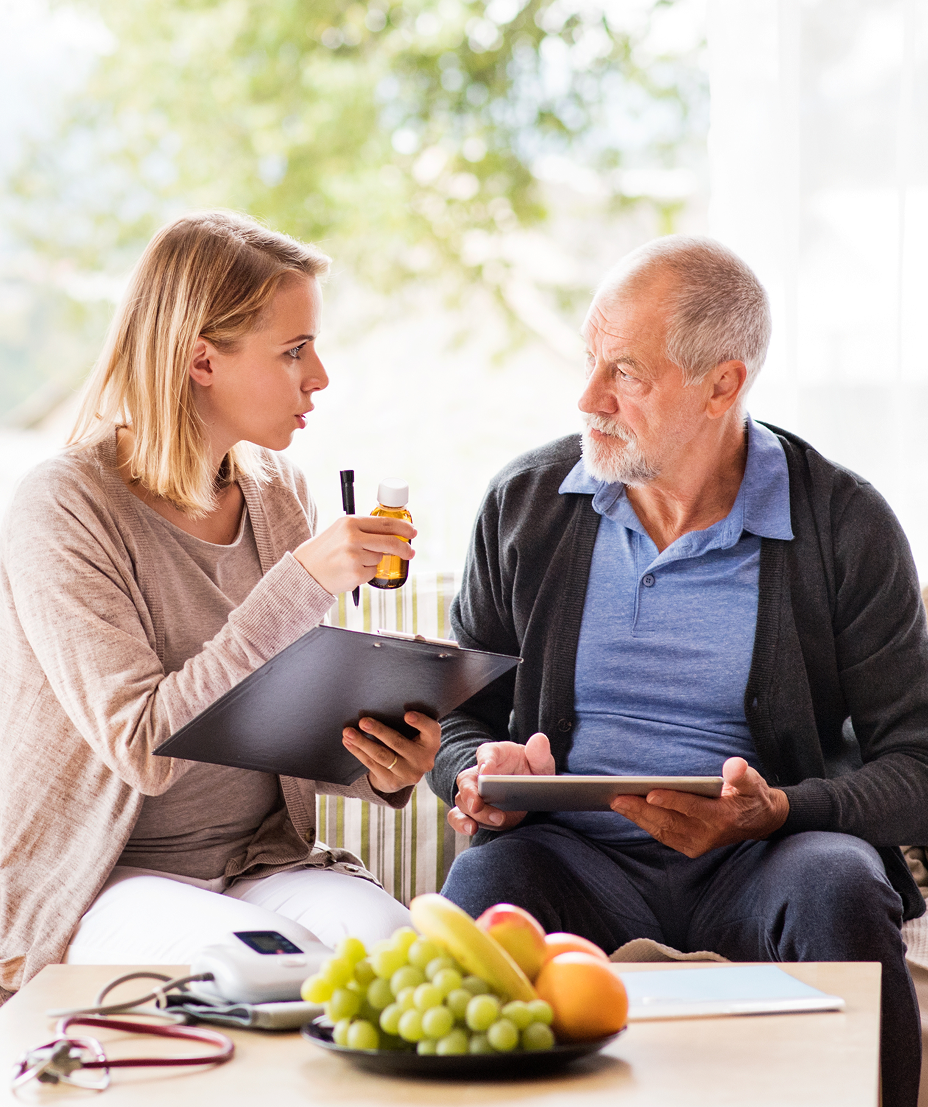 Jüngere Frau erklärt älterem Mann ein Medikamentfläschchen bei einem Beratungsgespräch im Wohnzimmer.