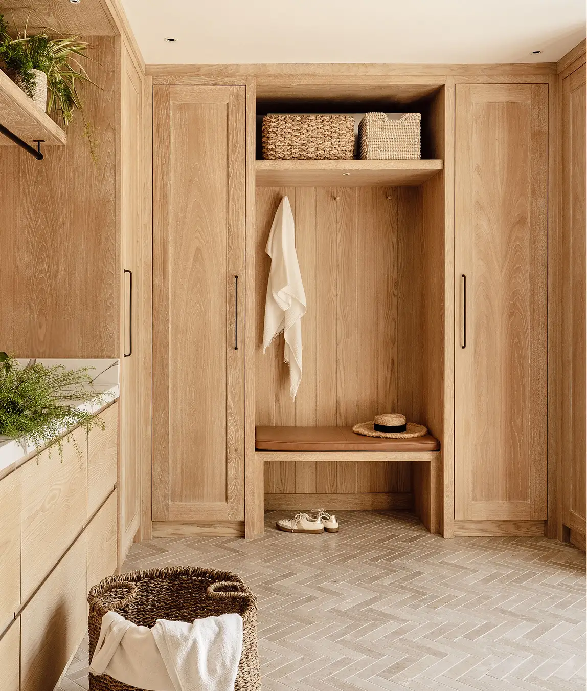 Light oak bootroom with built-in cabinets, woven baskets, a leather bench seat, and herringbone tile flooring.
