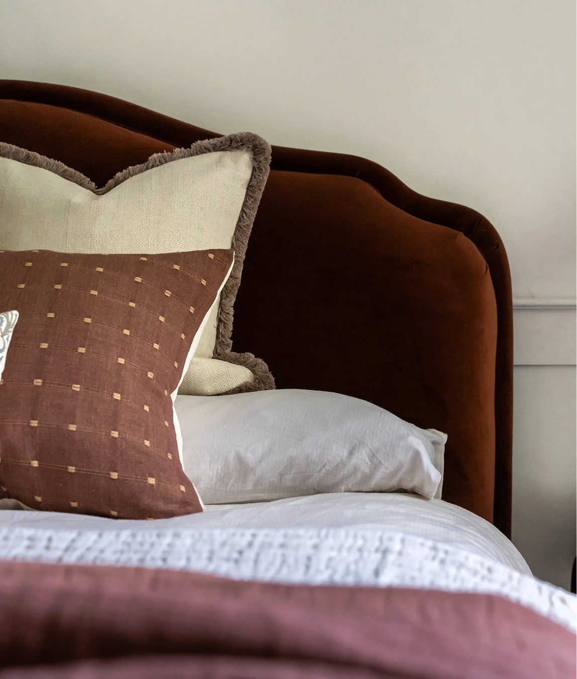 Cozy bed with rust velvet headboard, layered white bedding, and textured cushions in earthy brown and cream tones.