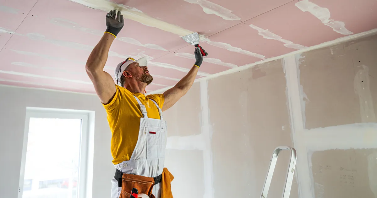 The worker makes a plasterboard ceiling. He does taping plaster drywall ceiling joints. A red plasterboard improve the fire resistance of ceiling structures.