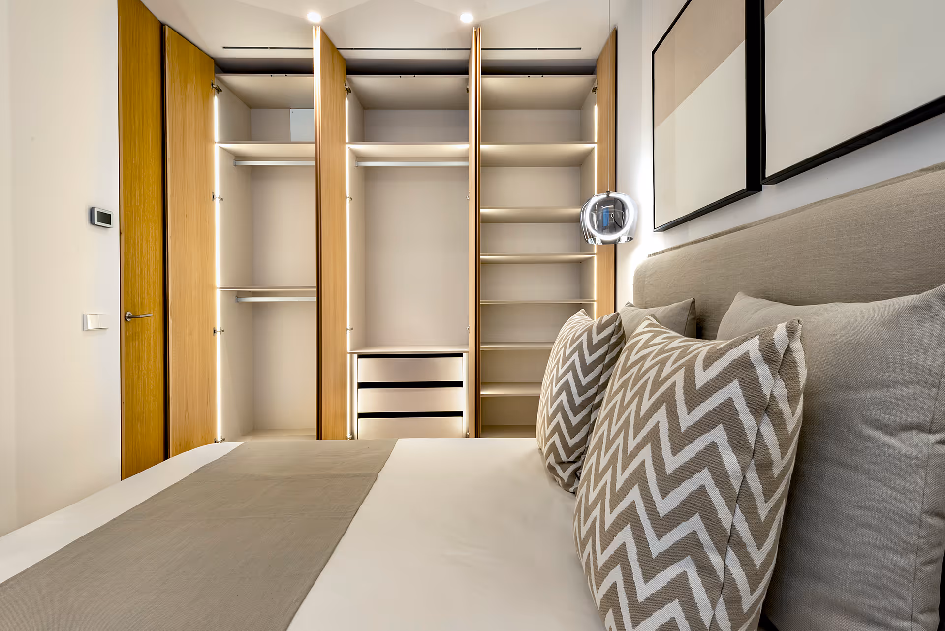 Modern bedroom with a neatly made bed featuring patterned pillows and an open wooden wardrobe with built-in lighting.
