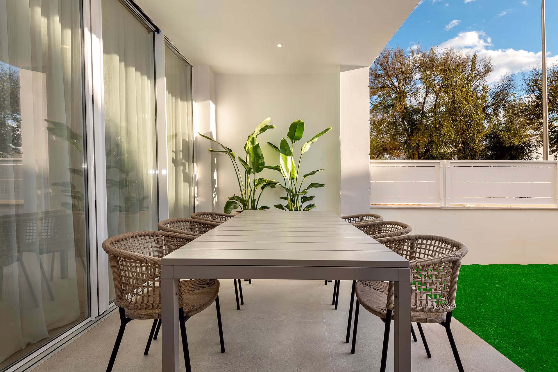 Outdoor patio dining area with a long rectangular table, six woven chairs, and potted green plants against a white wall.