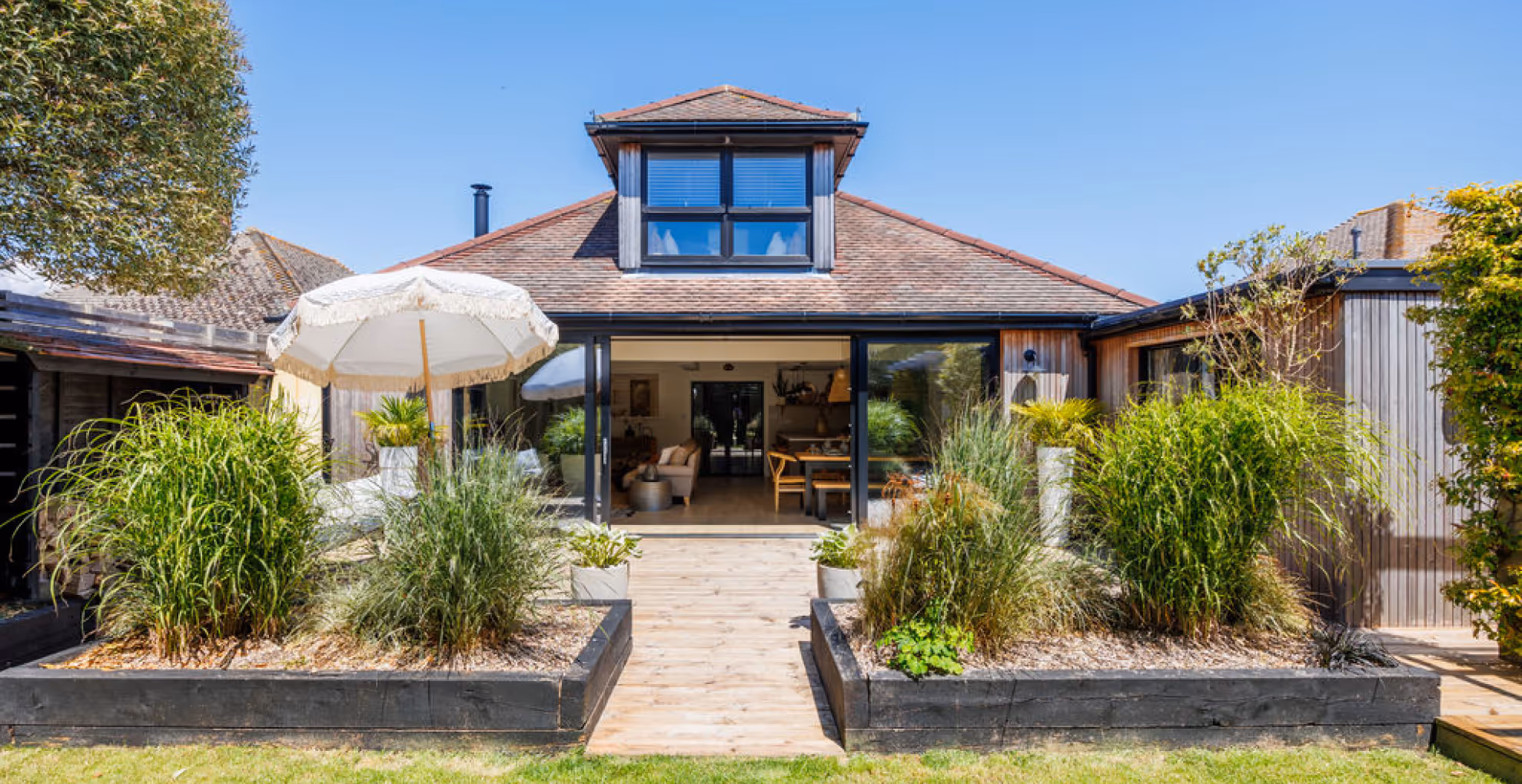 Modern house with sliding glass doors opening to a garden with plants, umbrella, and wooden pathway.