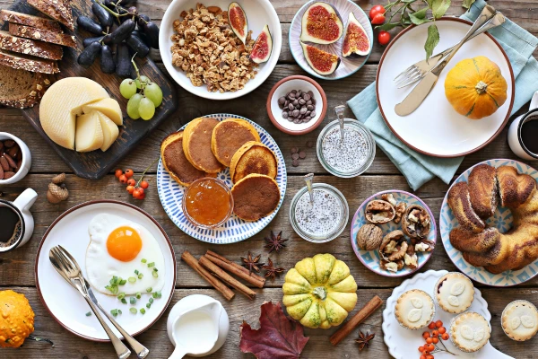 Herbstliches Frühstück mit Pfannkuchen, Spiegelei, Käse, Granola, Feigen, Kürbissen, Nüssen und Kaffee auf Holztisch.
