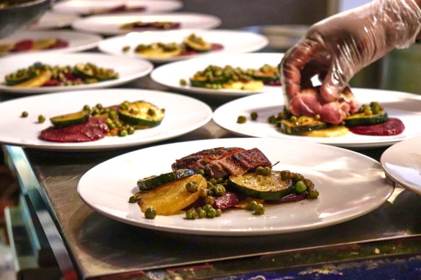 Ein Koch mit Handschuhen richtet auf mehreren weißen Tellern eine Vorspeise mit gegrillten Zucchinischeiben, Erbsen und einer kleinen Fleischportion an.