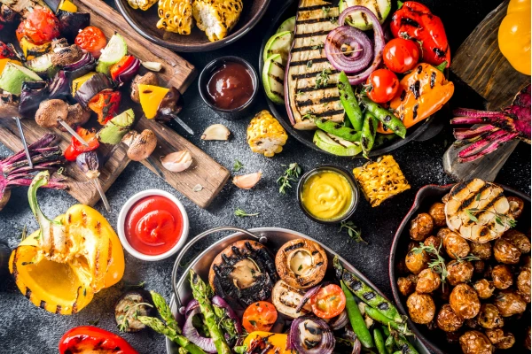 Bunte gegrillte Gemüseplatte mit Paprika, Pilzen, Mais, Tomaten und Soßen auf dunklem Tisch.
