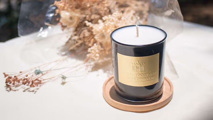 A stylish black candle with a gold label sitting on a wooden coaster, placed on a white surface. In the background, there is a wrapped bouquet of dried flowers, creating a rustic and natural atmosphere.