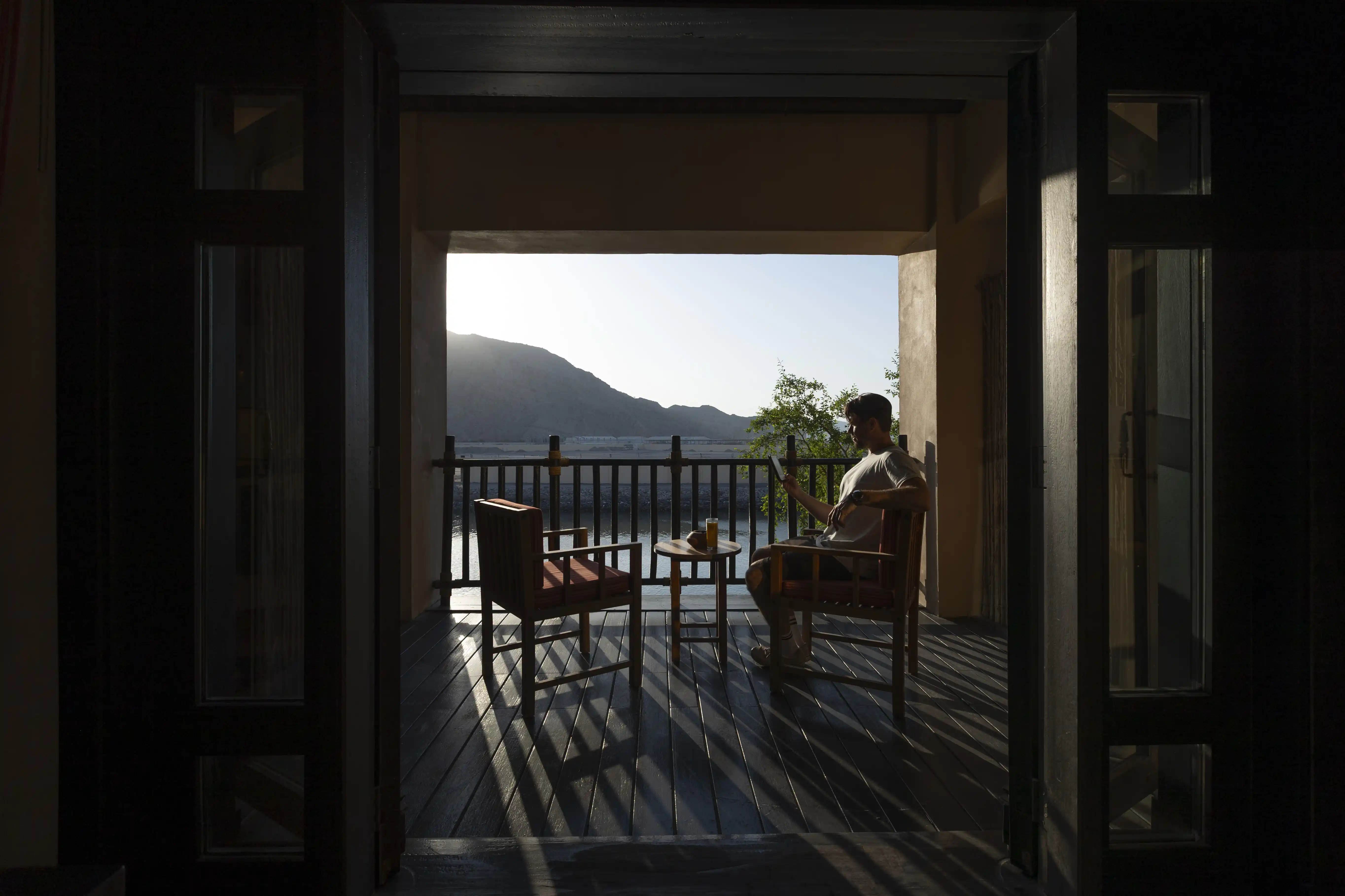 View from a darkened room framing a guest relaxing on the sunlit balcony, which overlooks the water and mountains.