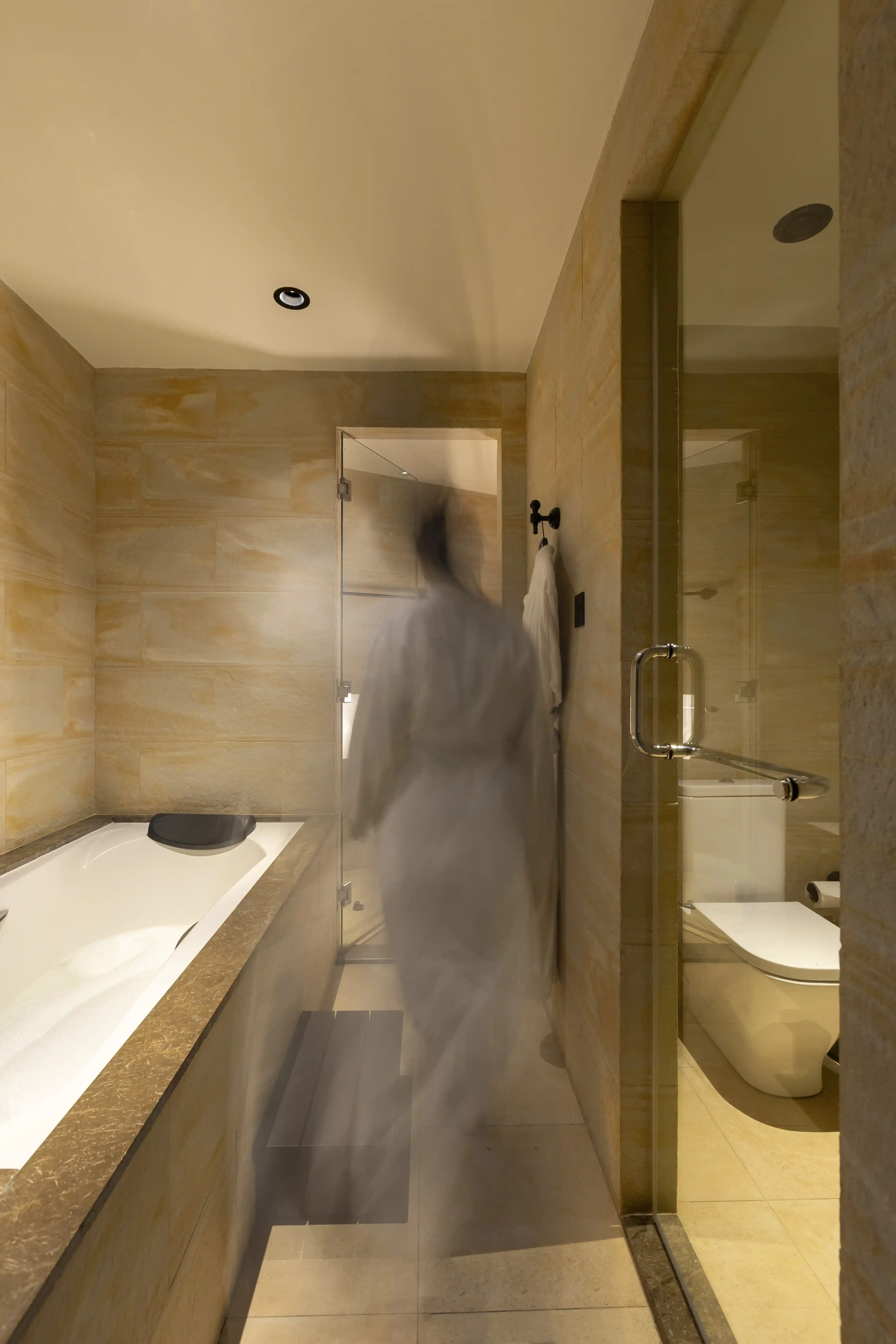 An artistic motion-blur shot of a guest in a white robe walking through the modern, tiled bathroom of the Musandam Suite.