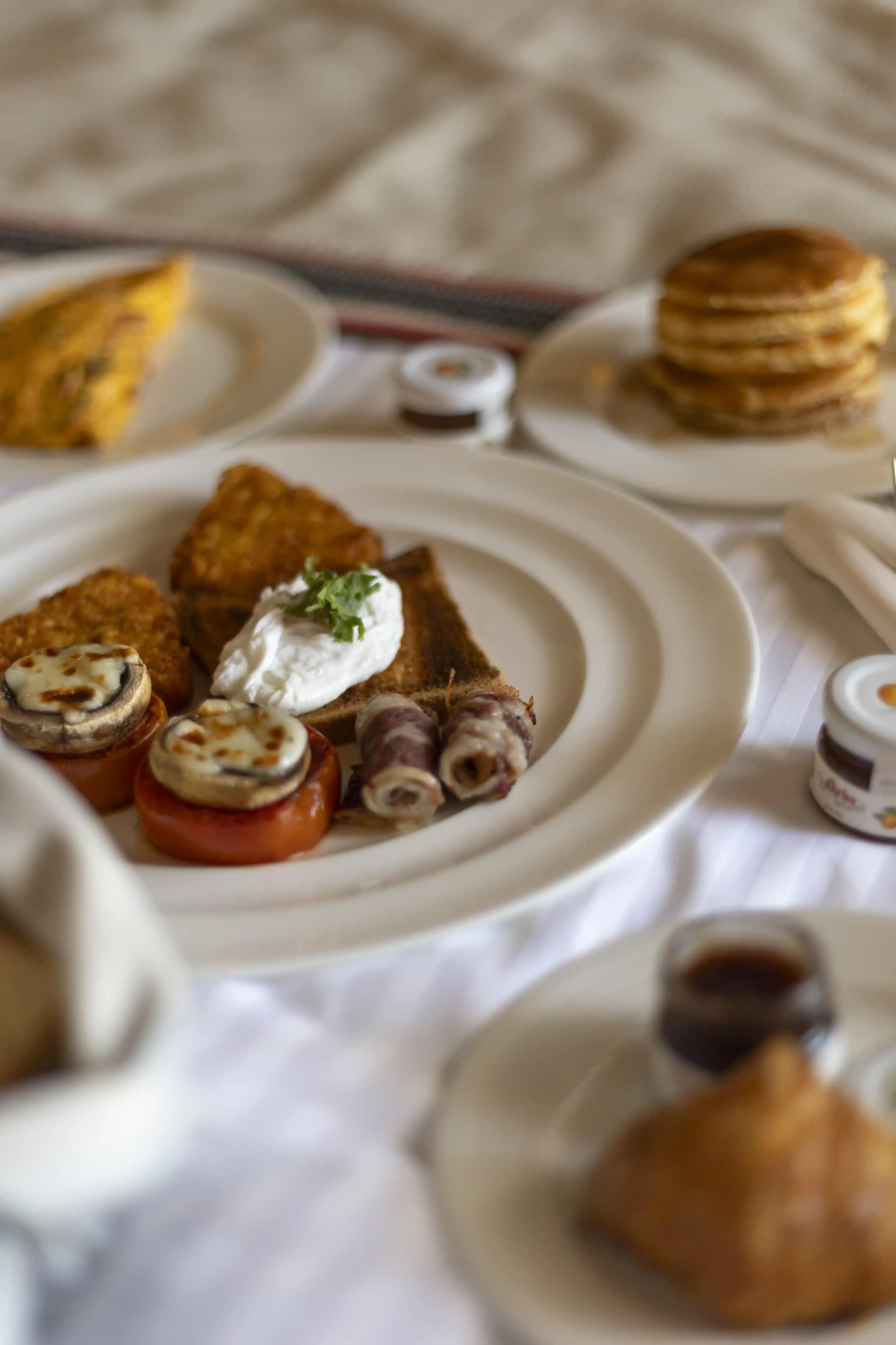 A close-up of a savory cooked breakfast plate featuring a poached egg on toast, hash browns, and grilled tomatoes, served in bed.