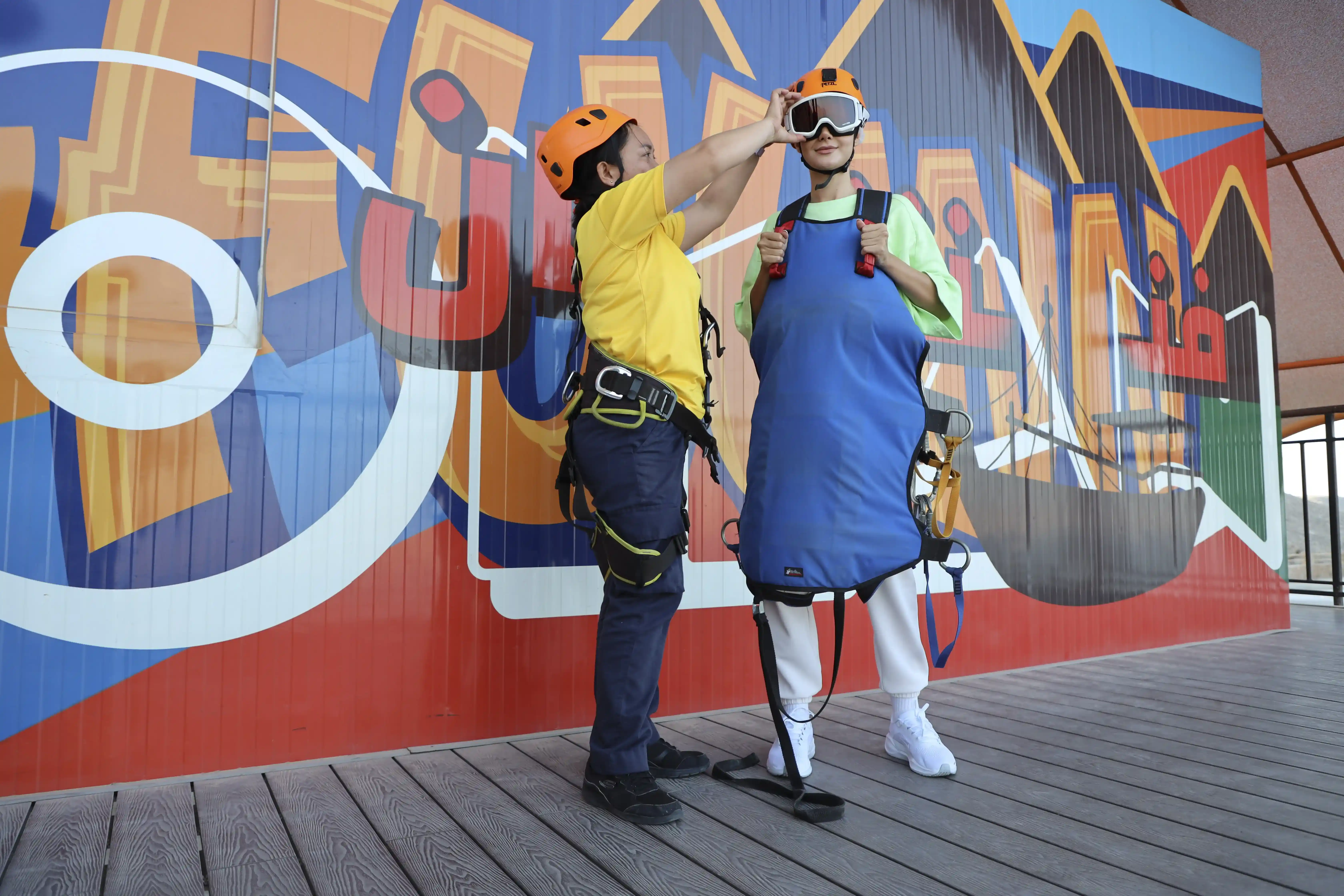An instructor helps a guest prepare for an adventure activity, adjusting their helmet and harness in front of a colorful mural.