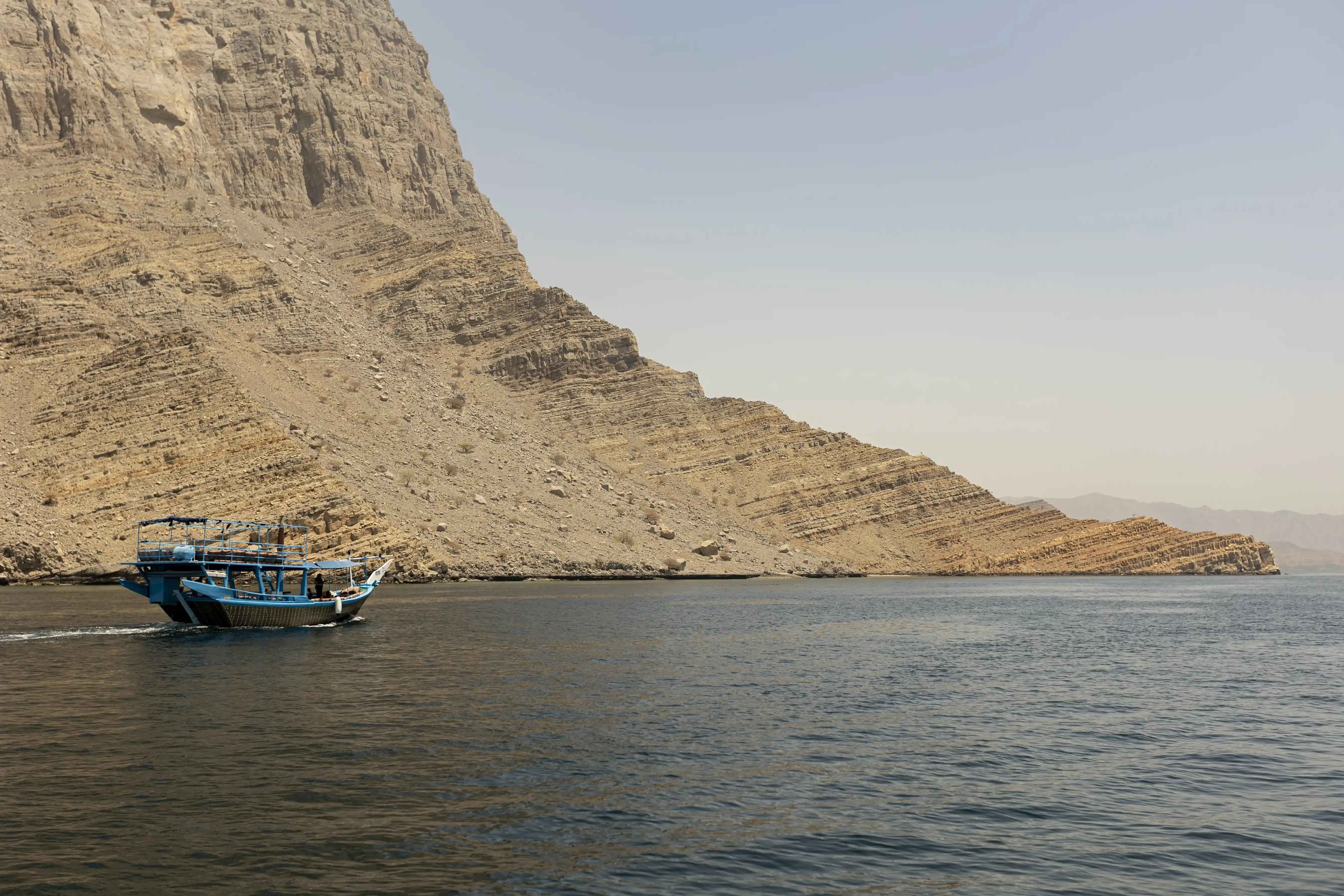 A blue dhow boat navigates the calm waters of a Musandam fjord, passing by a large, sloping rock formation.