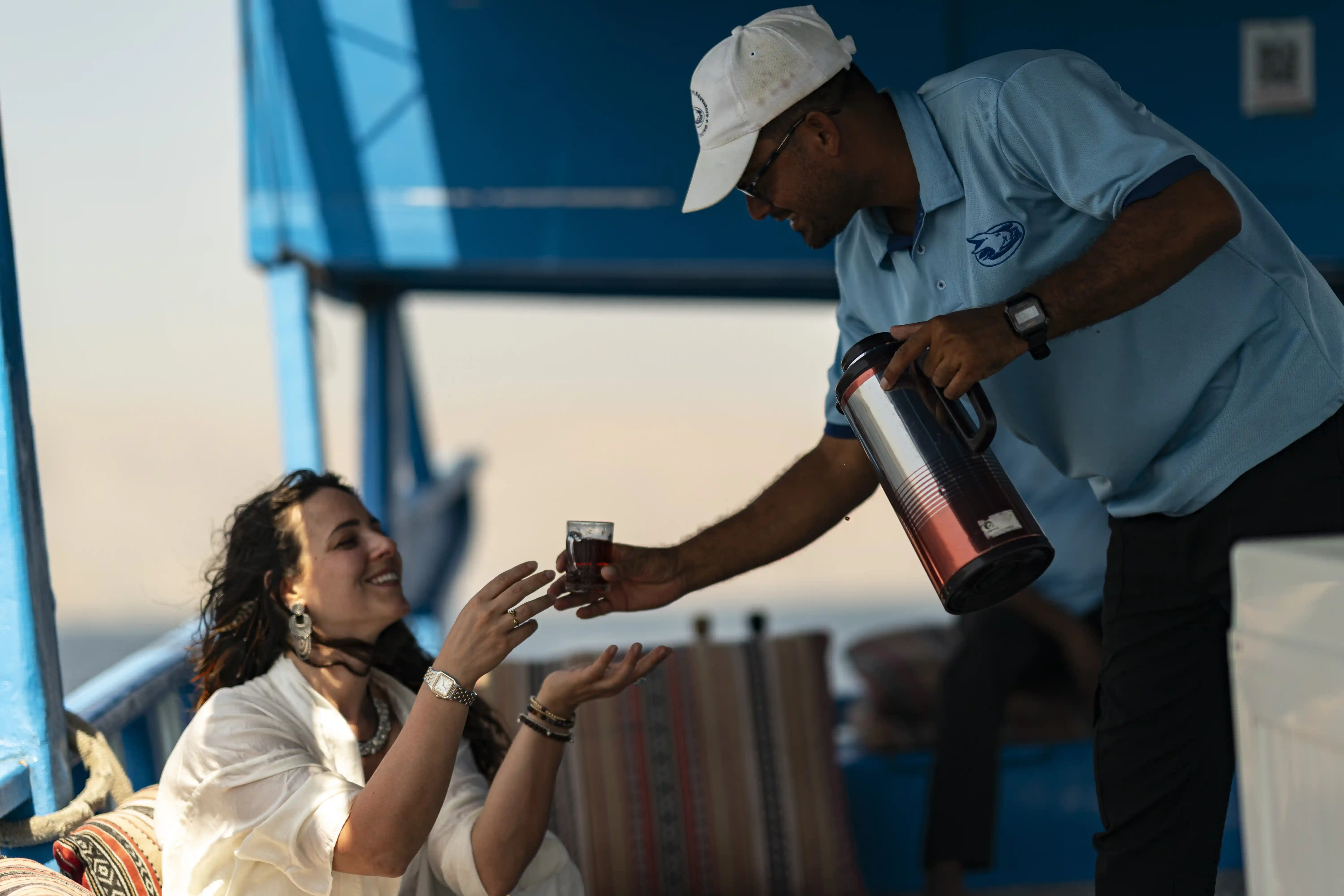 A crew member serves a welcome drink to a smiling guest aboard a traditional dhow boat during a cruise.