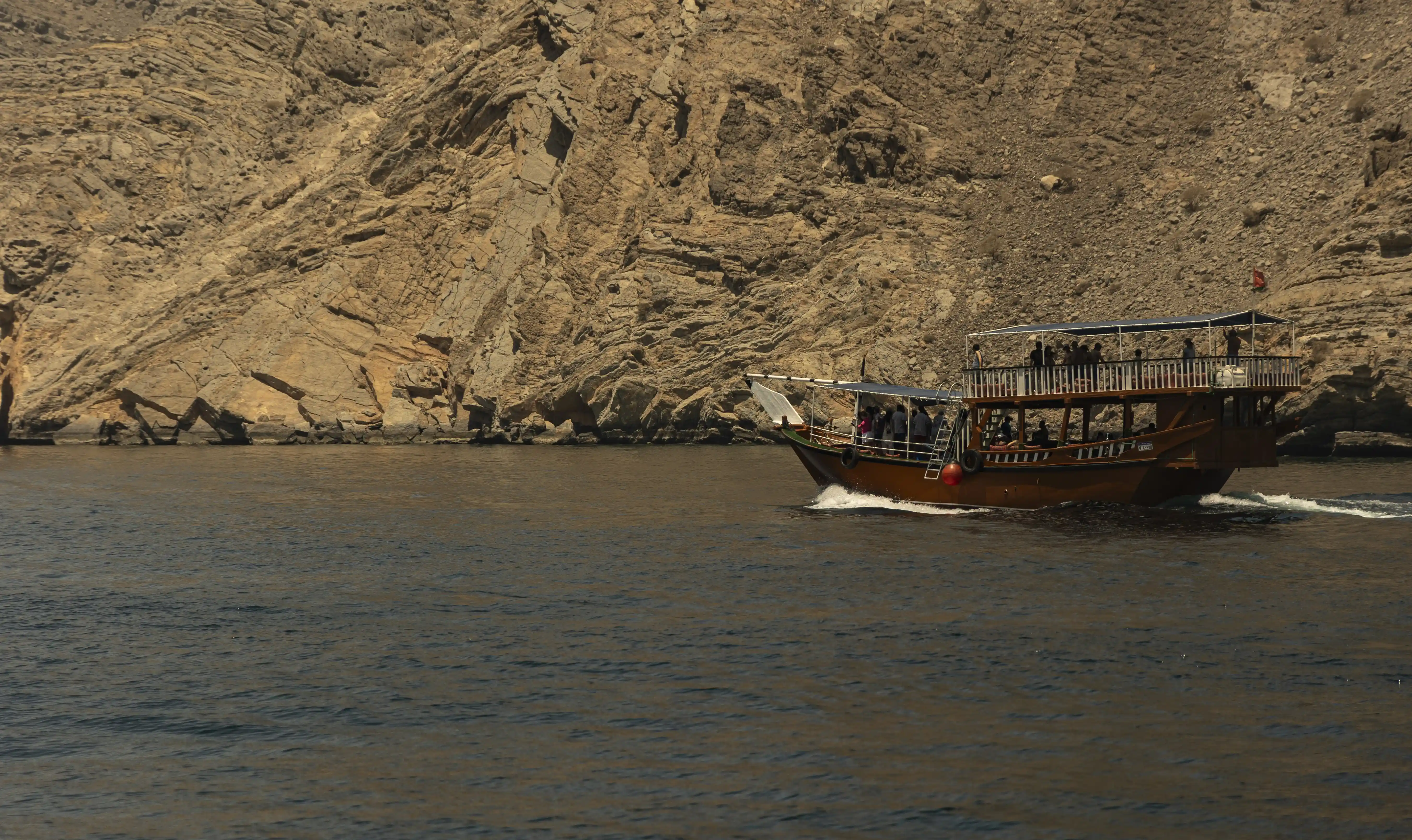 A traditional dhow boat filled with tour guests cruises alongside the immense, rocky cliffs of the Musandam fjords.