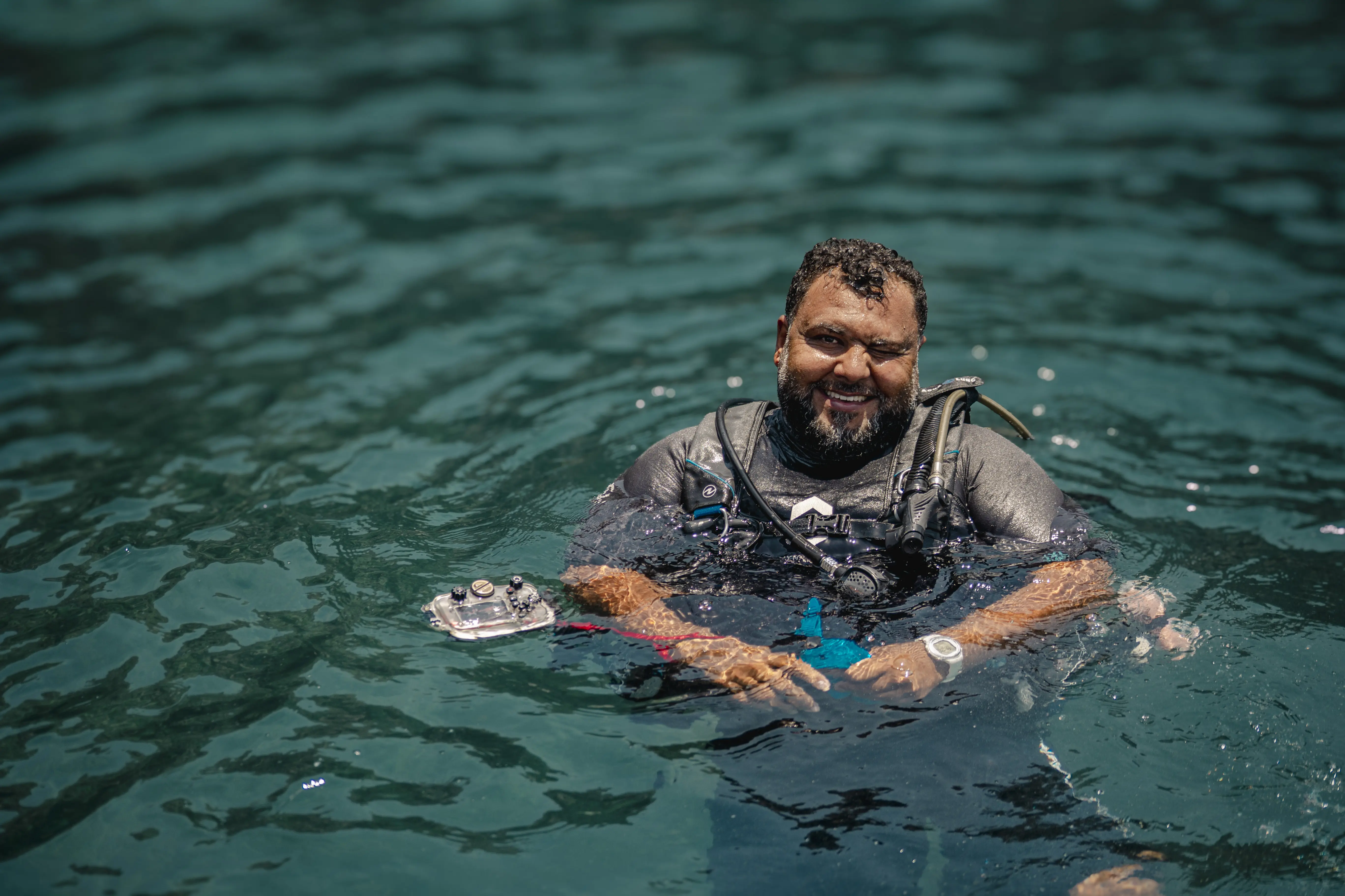 A happy scuba diver smiles after surfacing, holding an underwater camera in the clear water.