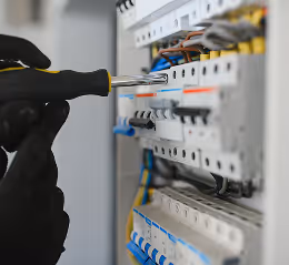 Hand in schwarzem Handschuh verwendet Schraubendreher an einem elektrischen Sicherungskasten.