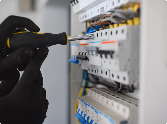 Hand mit schwarzem Handschuh, die Schraubendreher an einem Sicherungskasten benutzt.