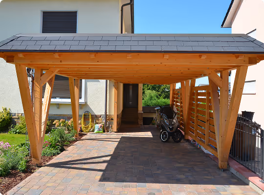 Holzcarport mit Schindeldach neben einem Haus, mit gepflastertem Boden und Kinderwagen auf der rechten Seite.