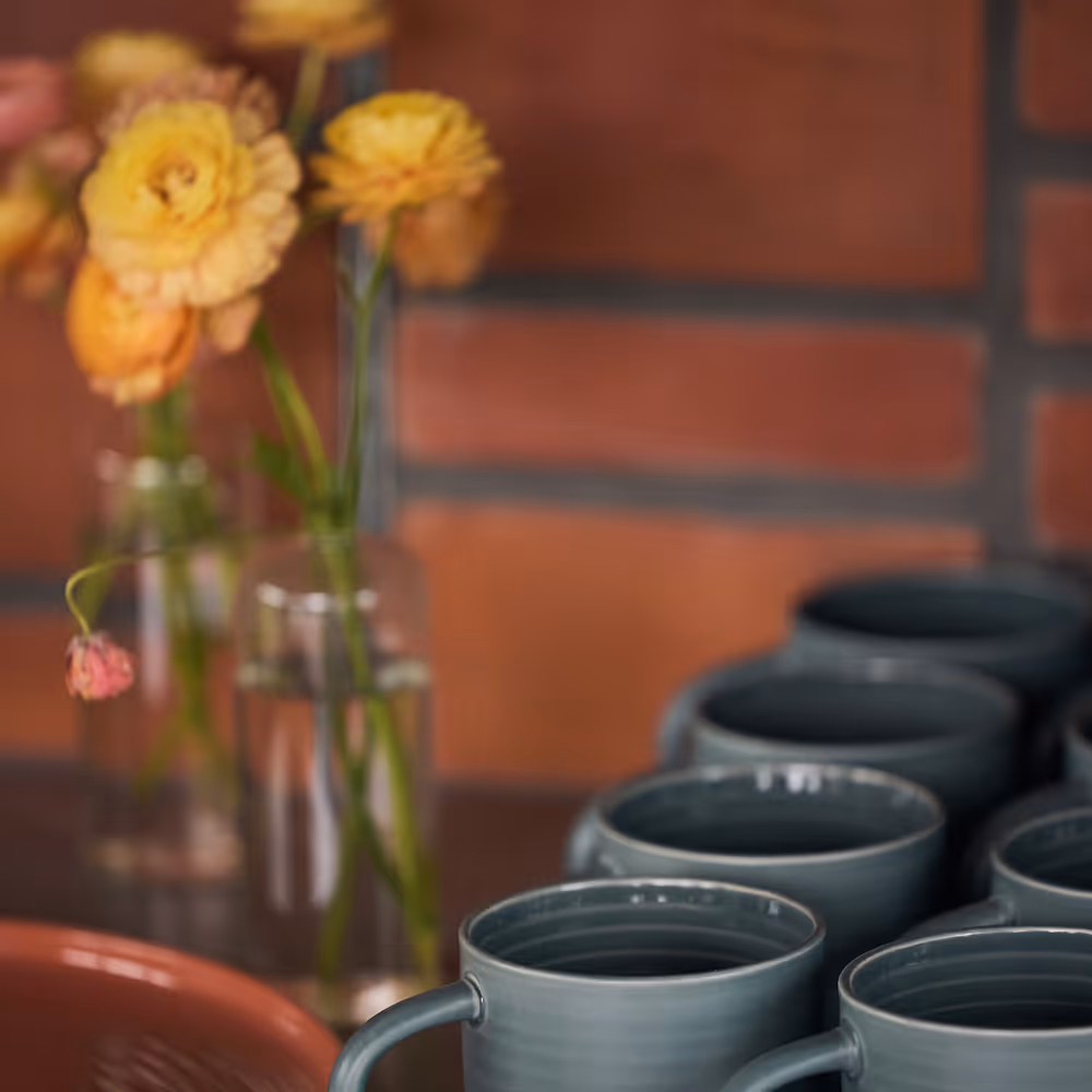 Vase of yellow flowers behind rows of blue grey mugs