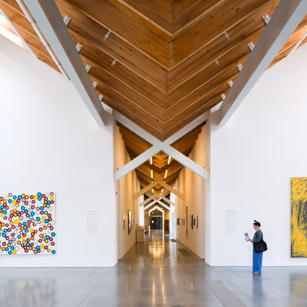 Person standing in museum with tall wood beam ceiling, a hallway in the middle and two paintings on the walls to either side of the hallway