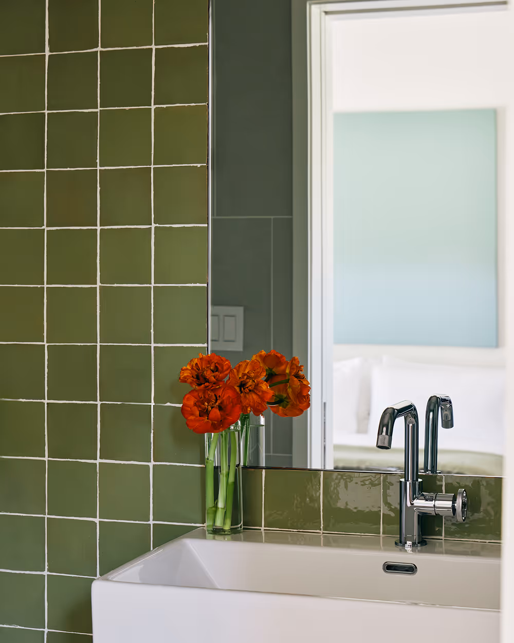 Green tile wall in bathroom with a mirror and red flowers in a vase on the sink edge