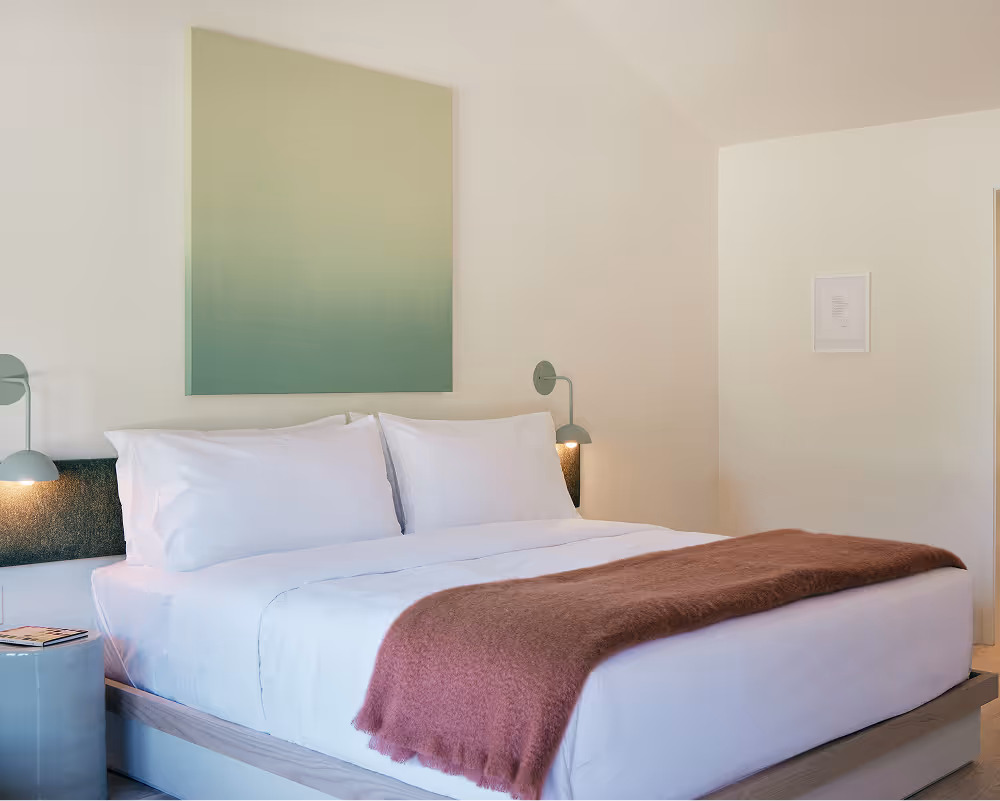 Angled view of king bed, side tables and sconces on either side, green painting above bed