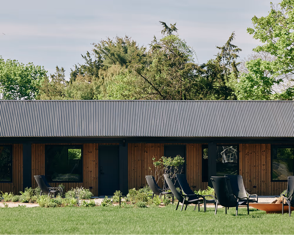 Wood panel hotel building with black accents, grass lawn with chairs and fire pit in front and and trees behind