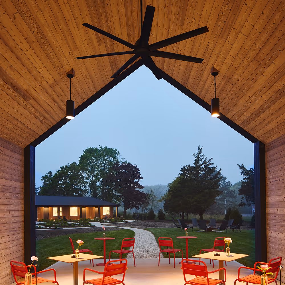Internal view of wood paneled barn with red dining sets looking out to a walkway lined with grass lawns leading to lit up hotel building