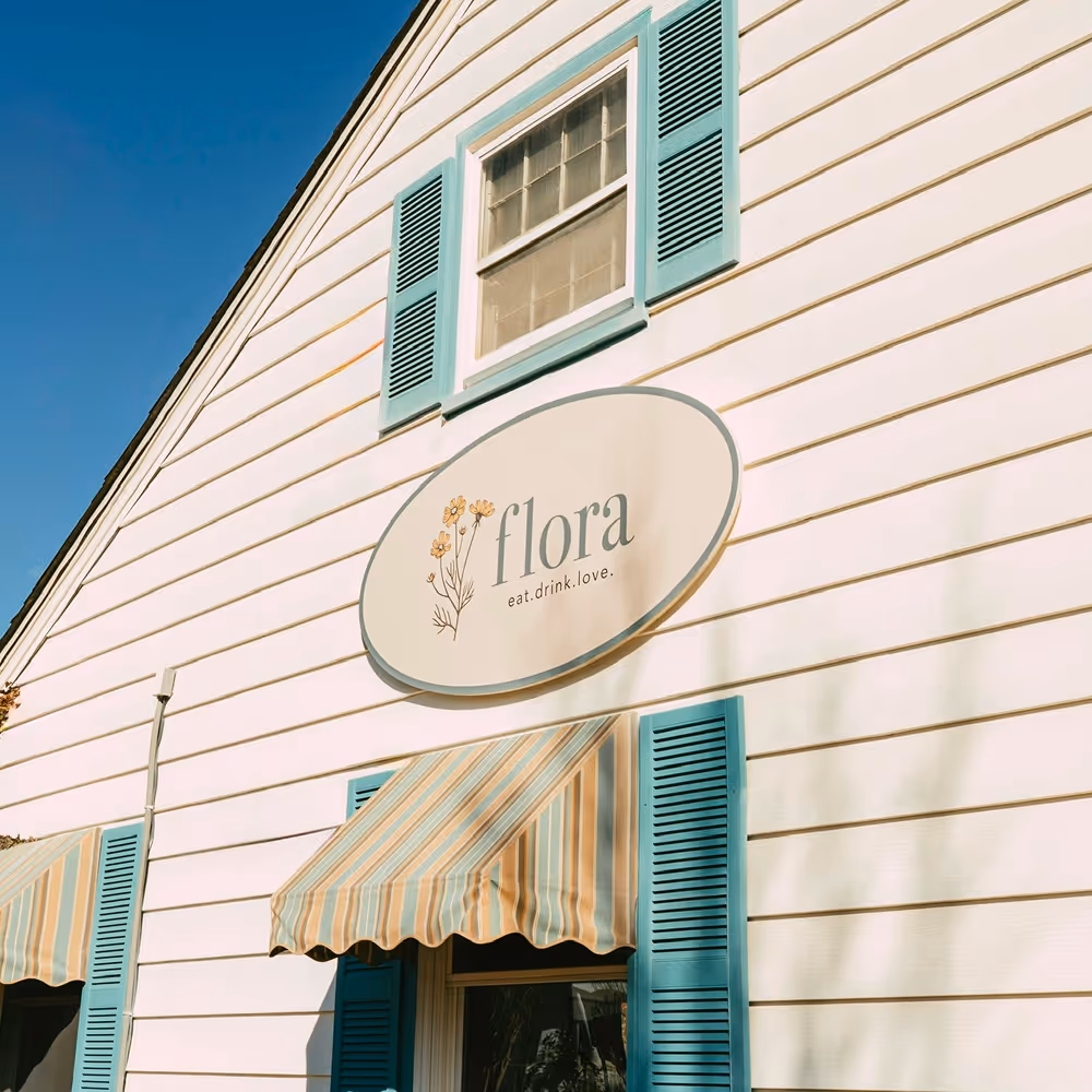 Oval sign that reads "flora eat. drink. love." on a white panel exterior of building with teal shutters