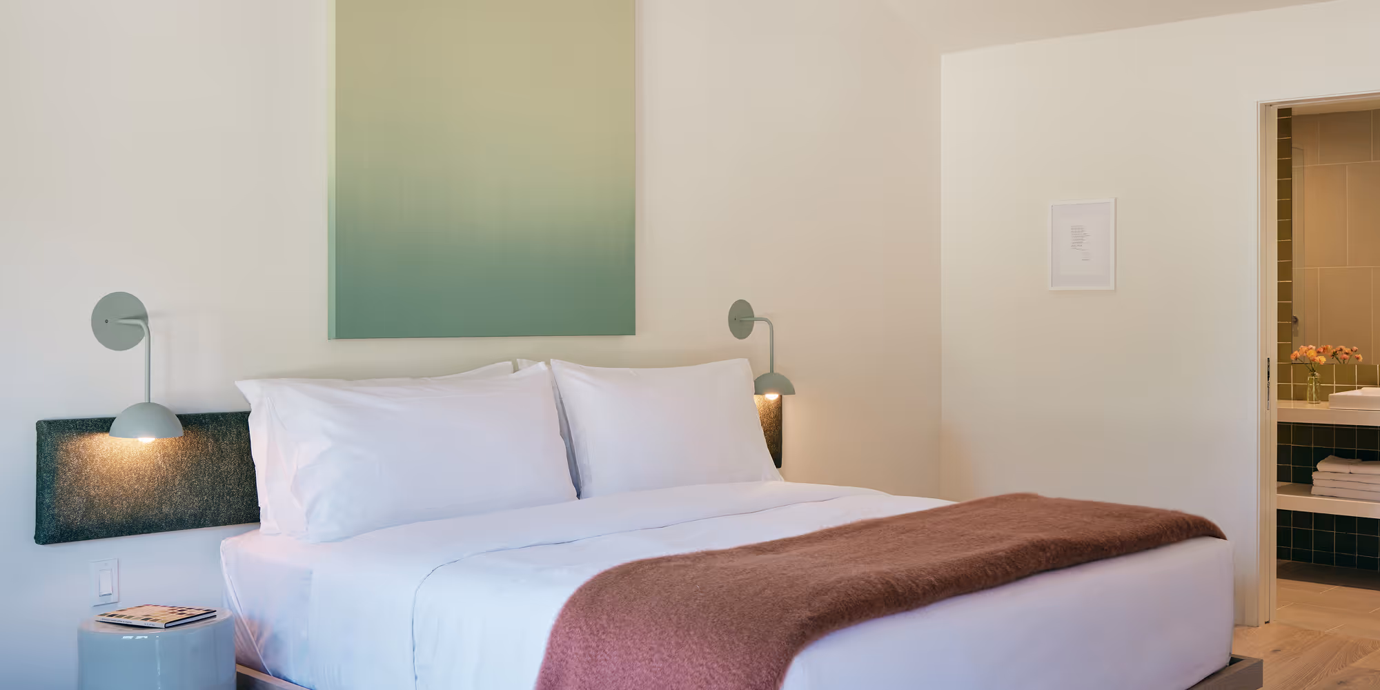 Angled view of king bed, side tables and sconces on either side, green painting above bed
