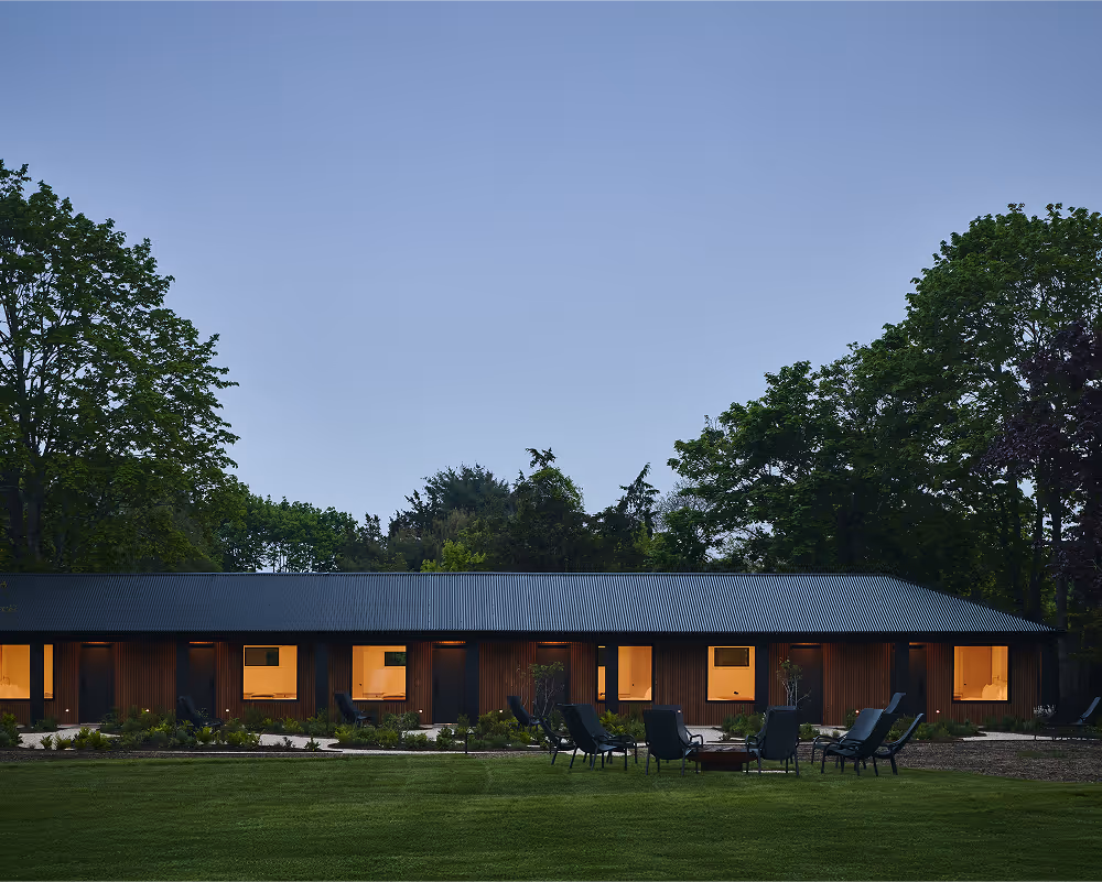 Wood panel hotel building with black accents, grass lawn with chairs and fire pit in front and and trees behind at sunset