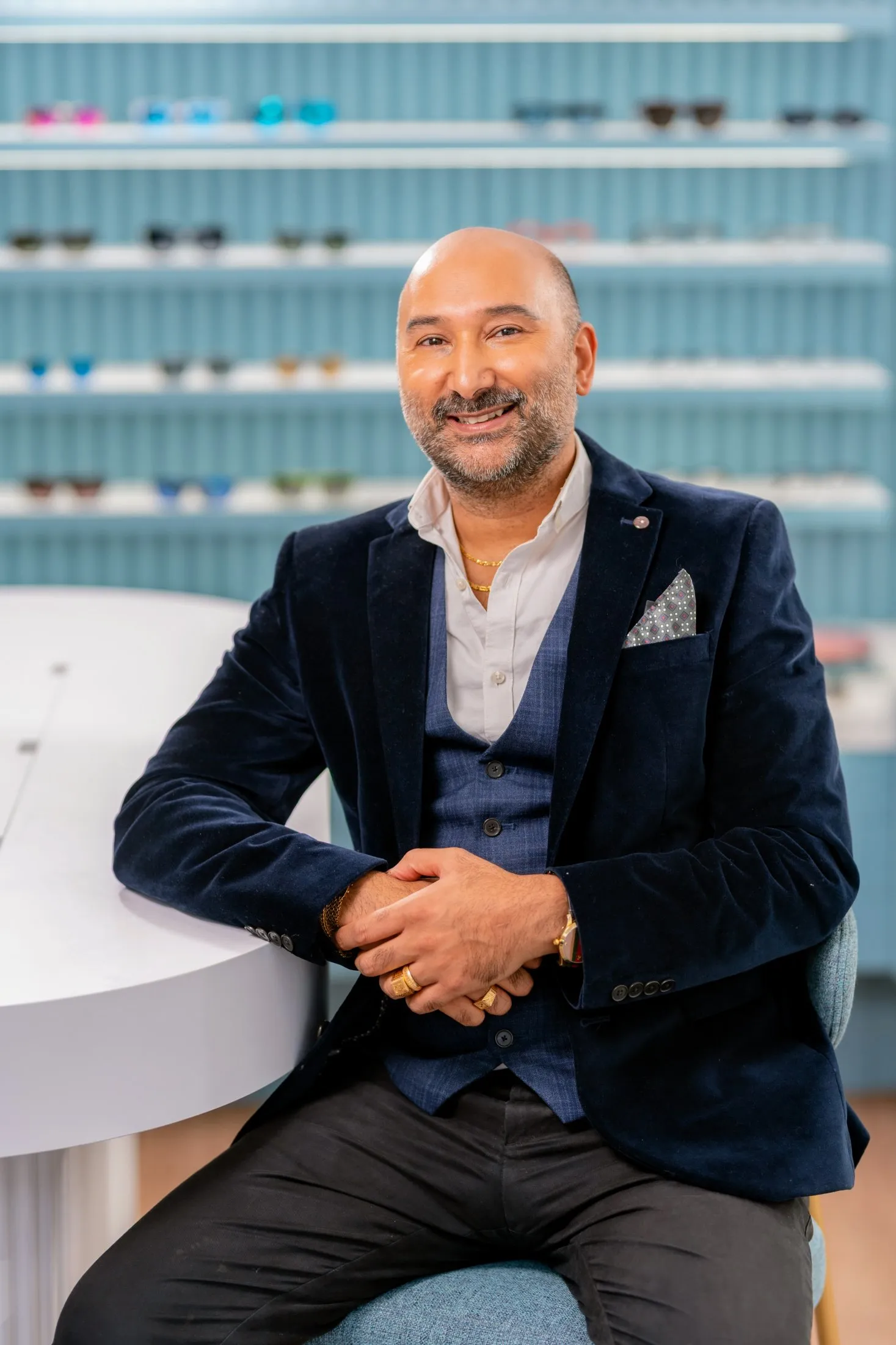 Mitesh Smiling professional in navy blazer sitting in optical store with glasses shelves