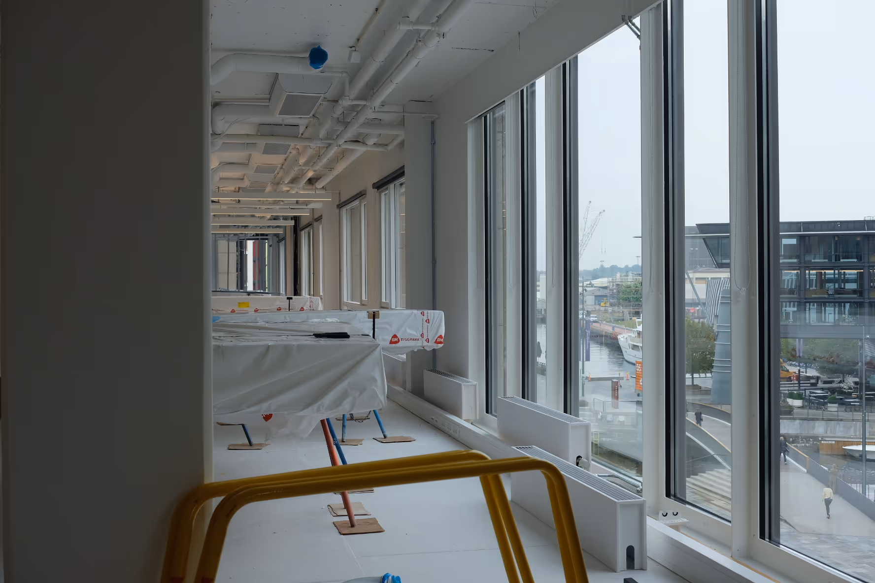 Kontorlokale under innredning med store gulv-til-tak-vinduer som gir utsikt mot en havnepromenade. Rommet er lyst og preget av pågående arbeid, med møbler dekket i plast og vernegjerder plassert i rommet.