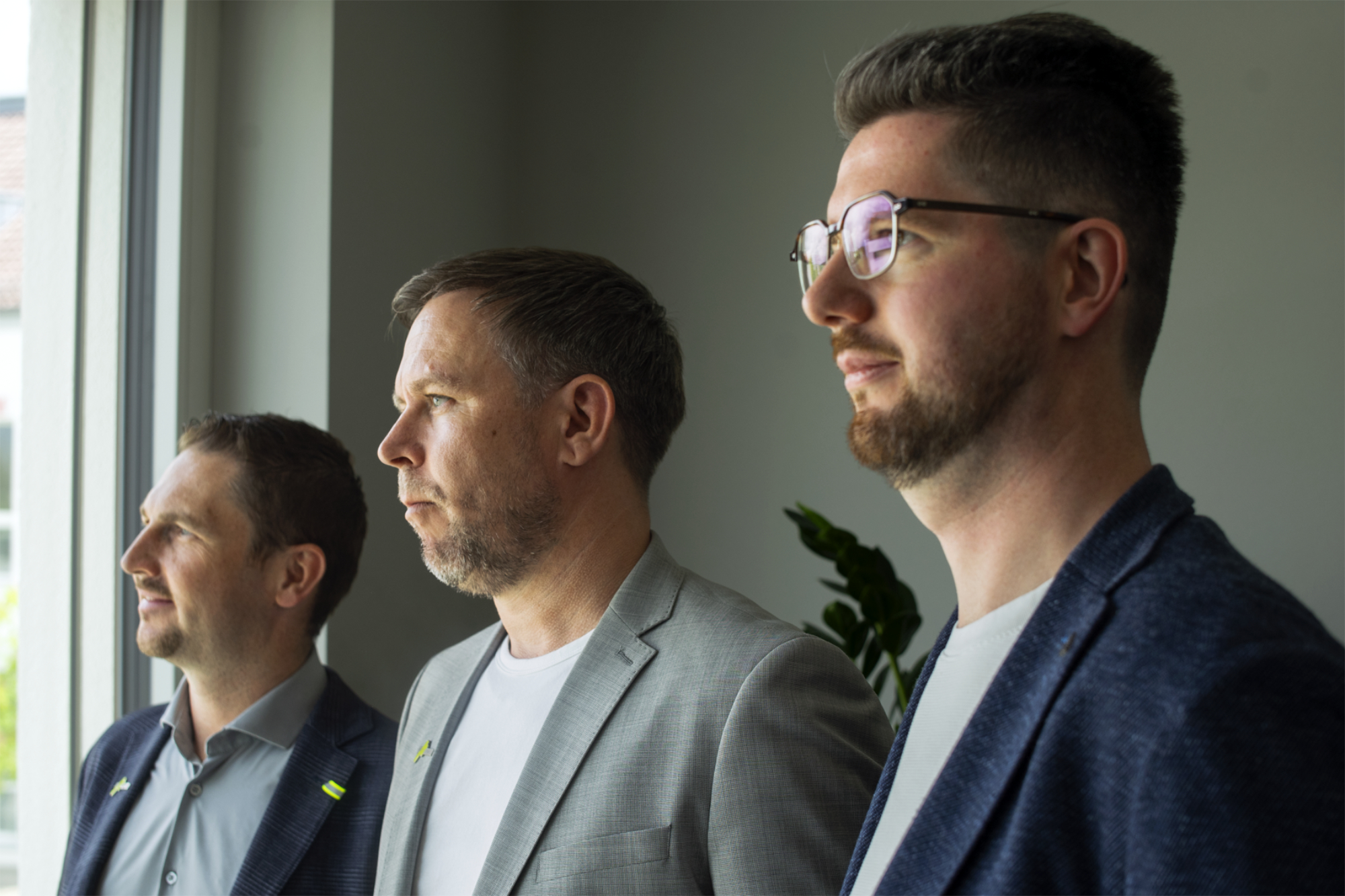 Drei Männer in Anzügen blicken nachdenklich aus einem Fenster in einem Büro.