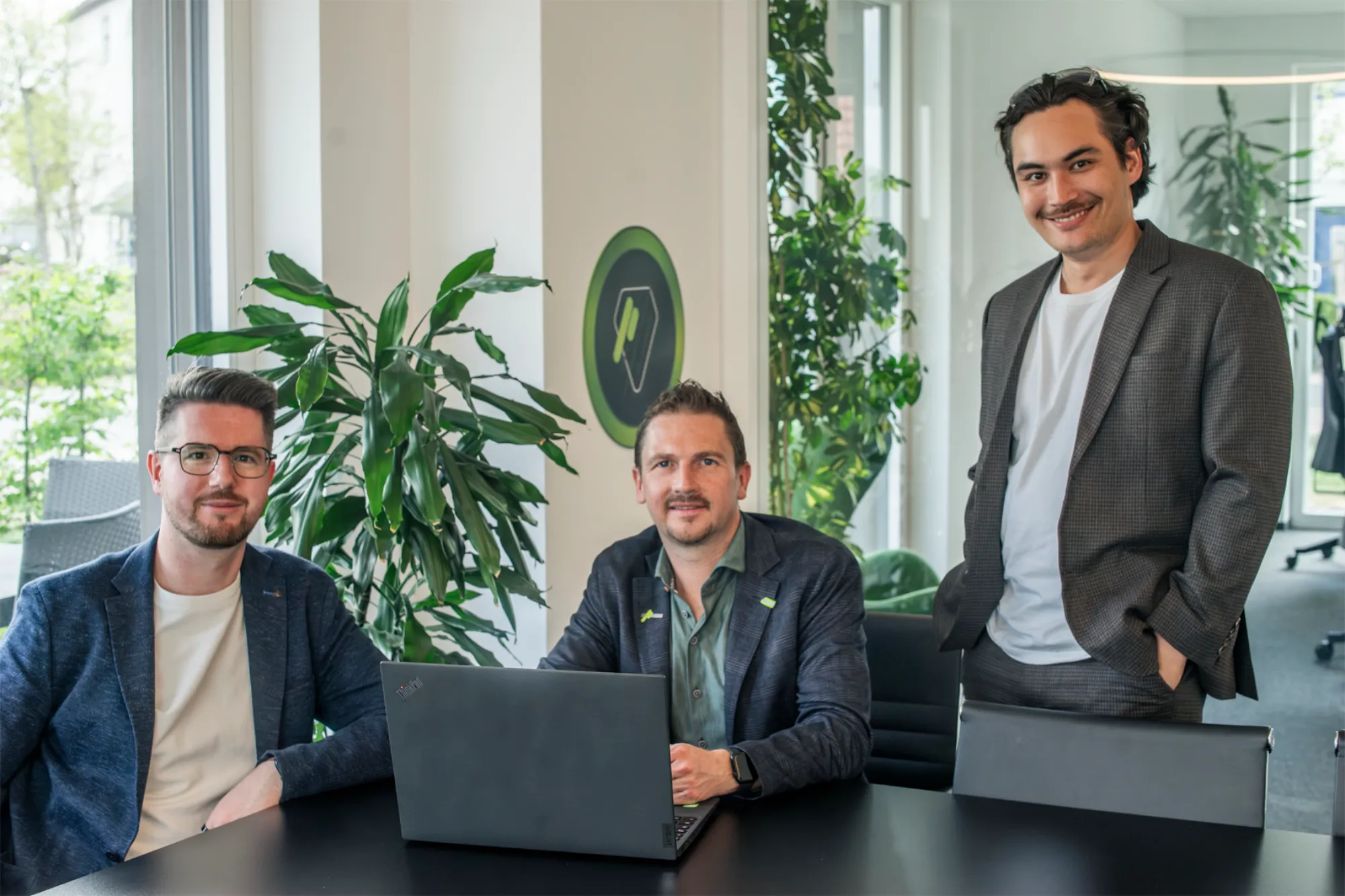 Drei Männer in lässiger Geschäftskleidung sitzen und stehen um einen Tisch mit Laptop in einem modernen Büro mit Pflanzen.