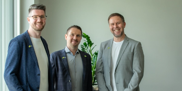 Drei Männer in Anzügen stehen lächelnd vor einer hellen Wand mit einer grünen Pflanze im Hintergrund.