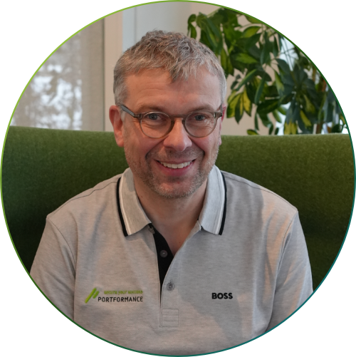 Ein lächelnder Mann mit Brille und grauem Haar, der ein graues Polohemd mit den Logos "PORTFORMANCE" und "BOSS" trägt, vor grünem Hintergrund und Pflanzen.