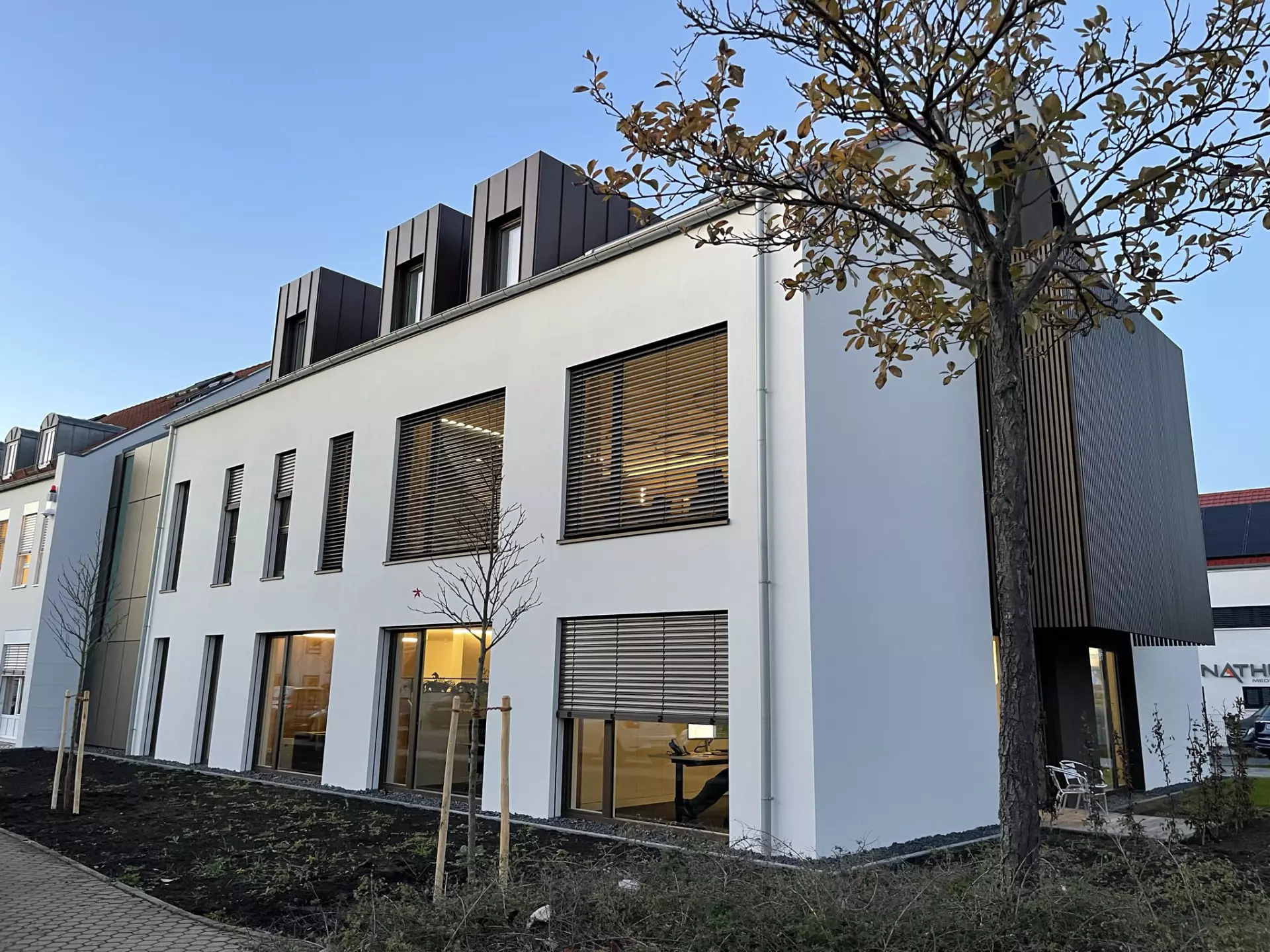 Modernes zweistöckiges Bürogebäude mit weißen Wänden, großen Fenstern und kleinen Bäumen im Vordergrund.