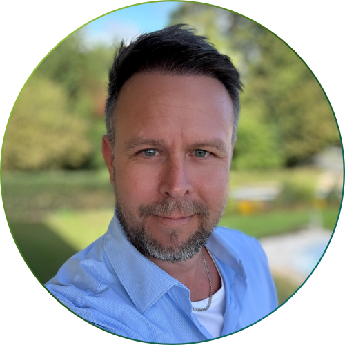 Nahaufnahme eines lächelnden Mannes mit kurzem braunem Haar und Bart, der ein hellblaues Hemd trägt, vor unscharf grünem Außenhintergrund.