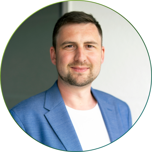 Mann mit kurzem braunem Haar und hellem Hemd trägt dunkelblauen karierten Blazer und lächelt in die Kamera.