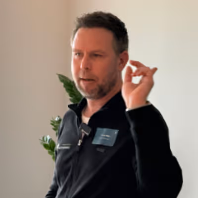 Mann mit Bart und dunkler Jacke spricht und hebt eine Hand mit leicht gespreizten Fingern.