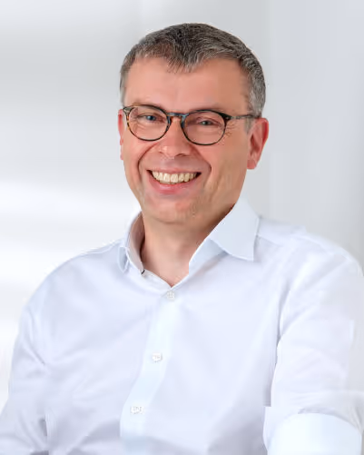 Lächelnder Mann mit kurzen braunen Haaren und Brille trägt weißes Hemd vor hellem Hintergrund.