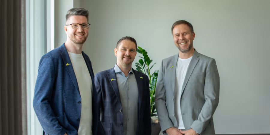 Drei Männer in Business-Kleidung lächeln vor einem neutralen Hintergrund mit einer Pflanze.