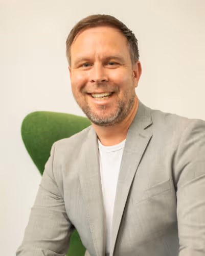 Lächelnder Mann in grauem Sakko vor hellgrünem Stuhl vor weißem Hintergrund.