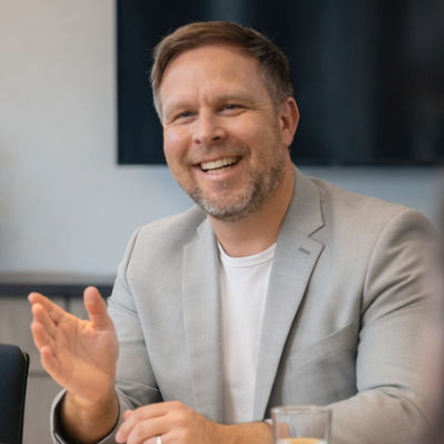 Mann mit Bart und dunkler Jacke spricht und hebt eine Hand mit leicht gespreizten Fingern.