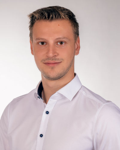 Porträt eines lächelnden Mannes mit kurzen braunen Haaren, in einem weißen Hemd und dunkelgrauem kariertem Sakko.