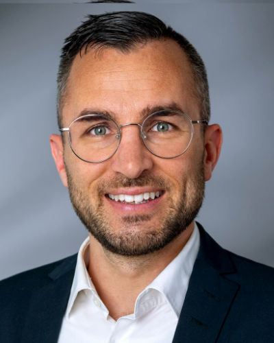 Porträt eines lächelnden Mannes mit kurzen braunen Haaren, in einem weißen Hemd und dunkelgrauem kariertem Sakko.